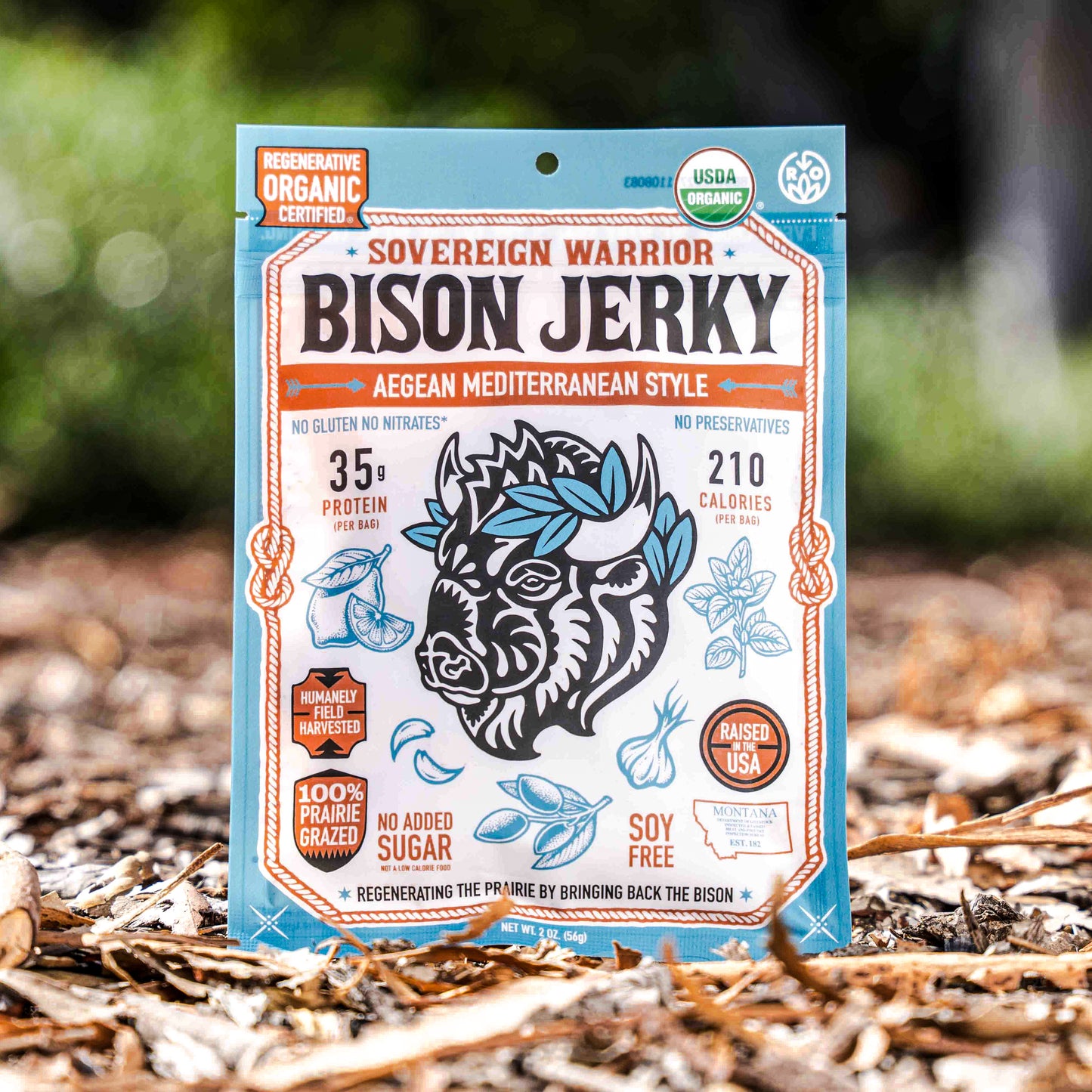 A bag of Sovereign Warrior Bison Jerky, AegEAN Mediterranean Style, standing upright on a bed of dry leaves and wood chips with a blurred forest background. The packaging features a light blue border, a central white panel with an illustration of a bison head decorated with blue leaves, and orange accents.