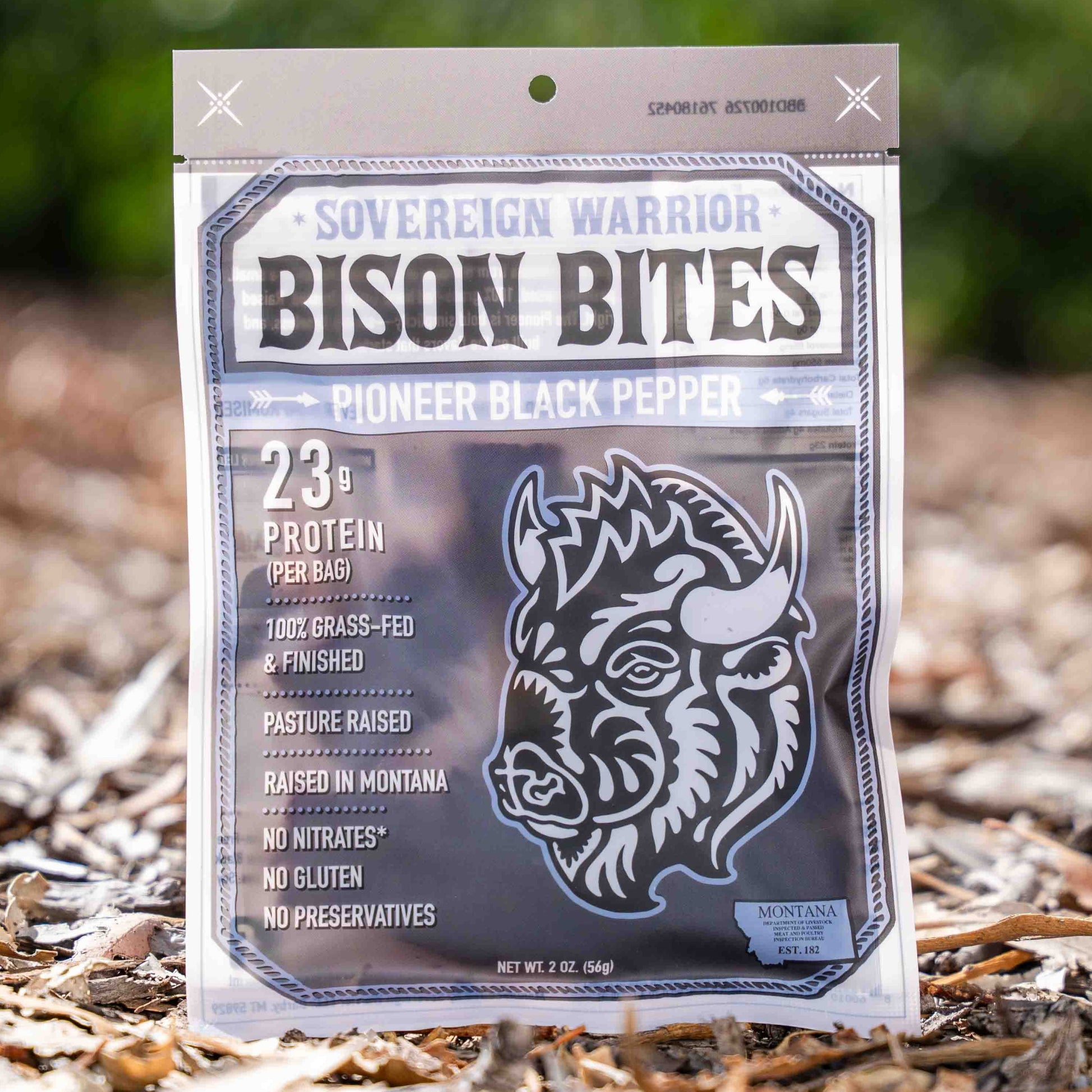 A bag of Sovereign Warrior Bison Bites 'Pioneer Black Pepper' protein treats standing upright on a bed of dry leaves and twigs outdoors, with a blurred green forest background.
