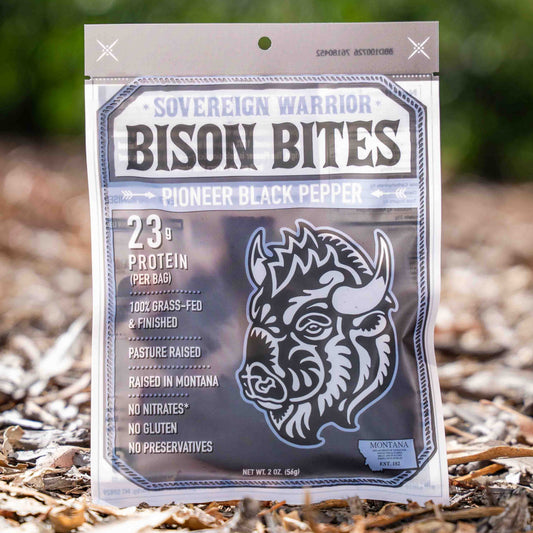 A bag of Sovereign Warrior Bison Bites 'Pioneer Black Pepper' protein treats standing upright on a bed of dry leaves and twigs outdoors, with a blurred green forest background.