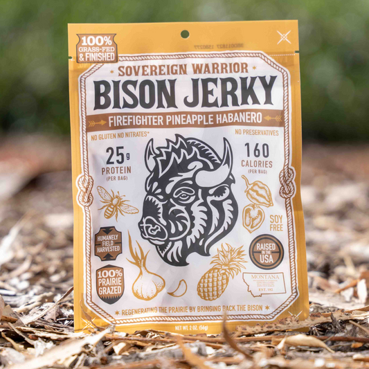 A bag of Bison Jerky, Firefighter Pineapple Habanero, standing upright on a bed of dry leaves and twigs with a blurred green forest background.