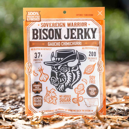 A bag of Sovereign Warrior Bison Jerky, Gaucho Chimurri flavor, standing upright on a bed of dried leaves and bark outdoors with a soft-focus green forest background.