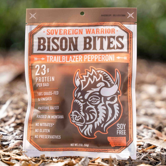 A bag of Sovereign Warrior Bison Bites Trailblazer Pepperoni dog treats stands upright on a bed of dry leaves and mulch outdoors, with a blurred green natural background.